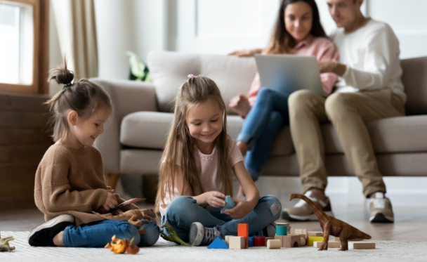Children playing with blocks and dinosaurs with parents on a couch in the background