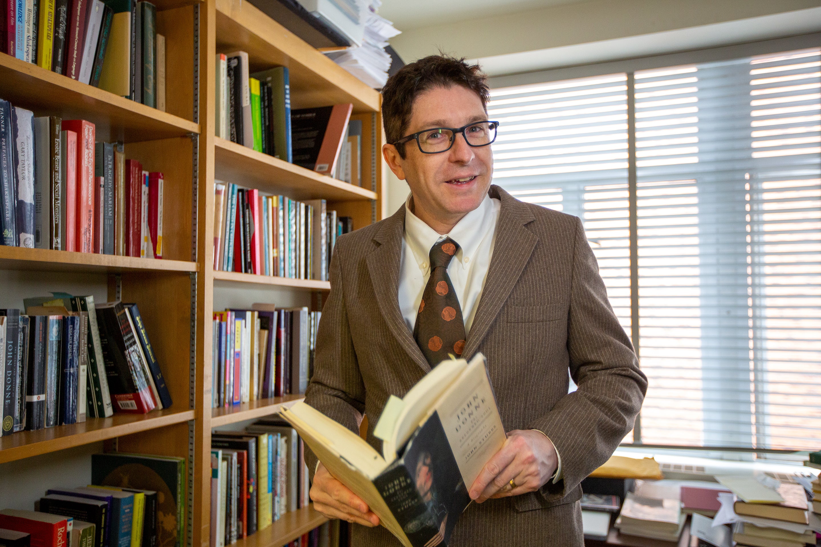 Professor Ben Saunders, standing in front of a bookshelf with a large book open in his hands