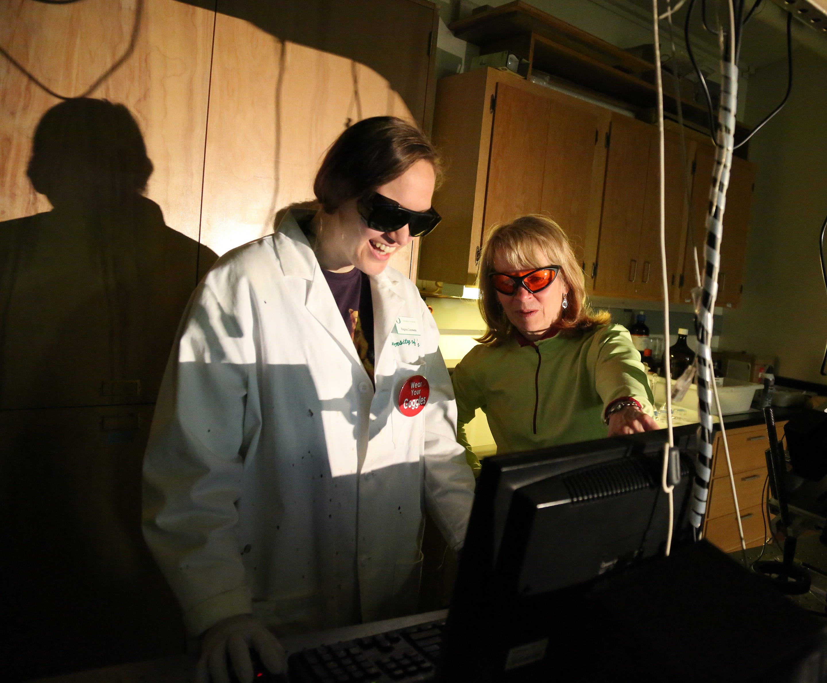 Prof. Geri Richmond in a light green sweater and orange safety glasses helping a student in a lab pointing to a computer monitor