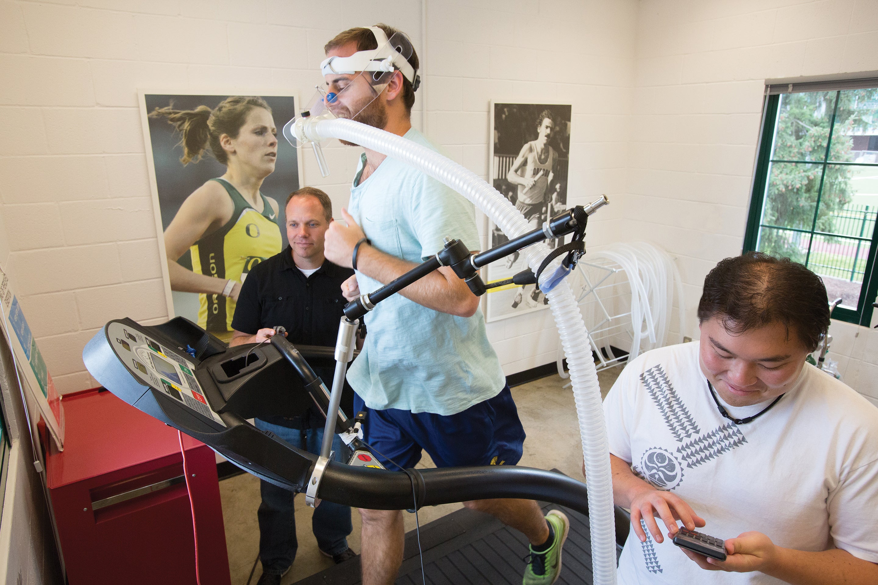 Athlete wearing a mask attached to a hose running on a treadmill with two people monitoring instruments