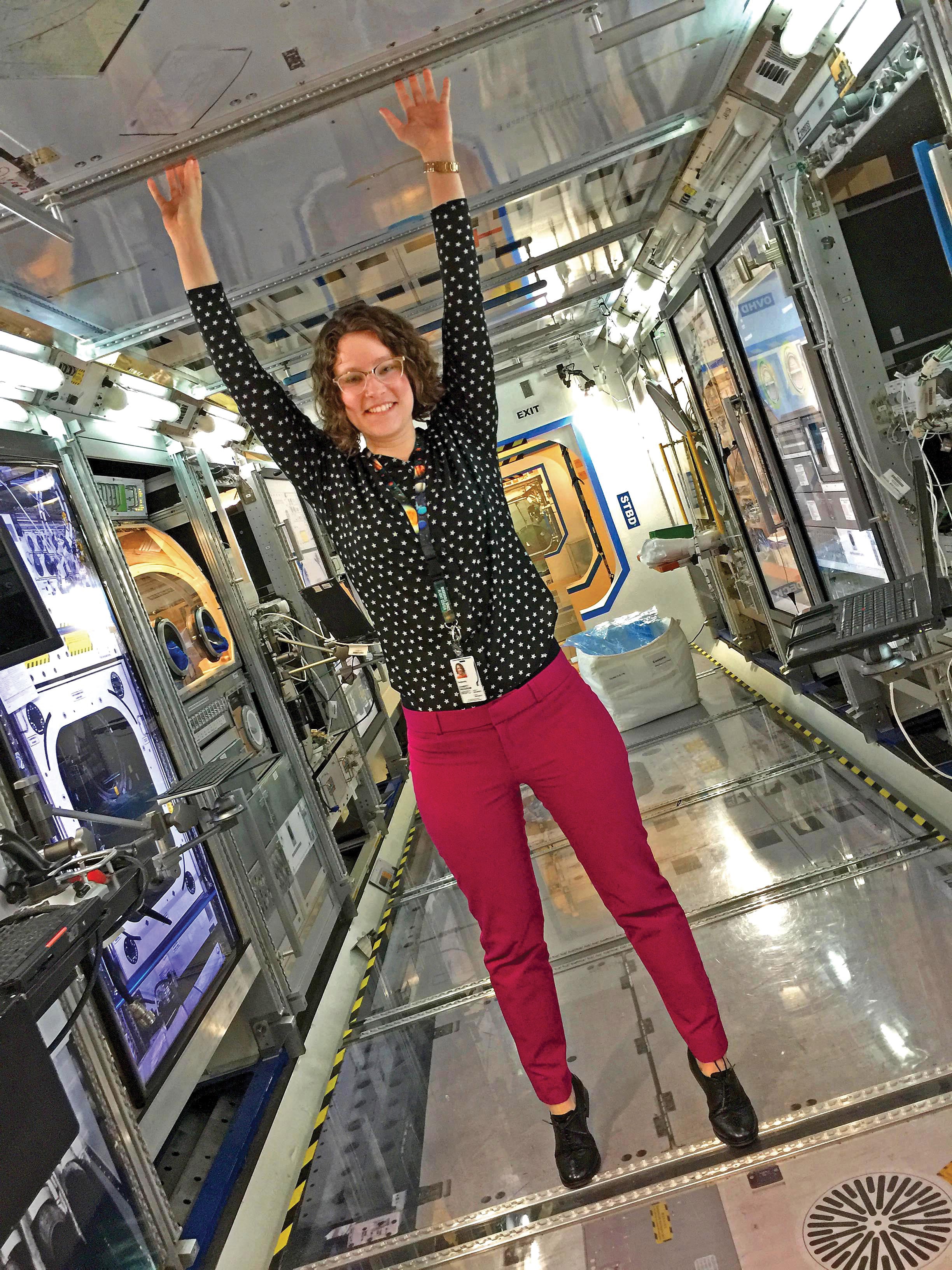 Charity Woodrum standing on tiptoes and touching the ceiling inside of a space-station laboratory mockup.