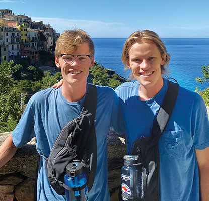 Riley Farrell and Bretten Farrell standing for a picture. Behind them, a stone wall, then a hillside town on the right, and the ocean on the left.