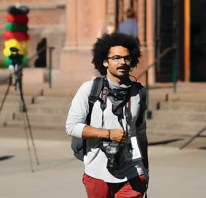 Jeremy Williams with a camera, zoom lens attached, journalist credentials, and a backpack walking in front of a sandstone building and a baloon banner showing red, yellow, green, and black colors of the Ghanian flag