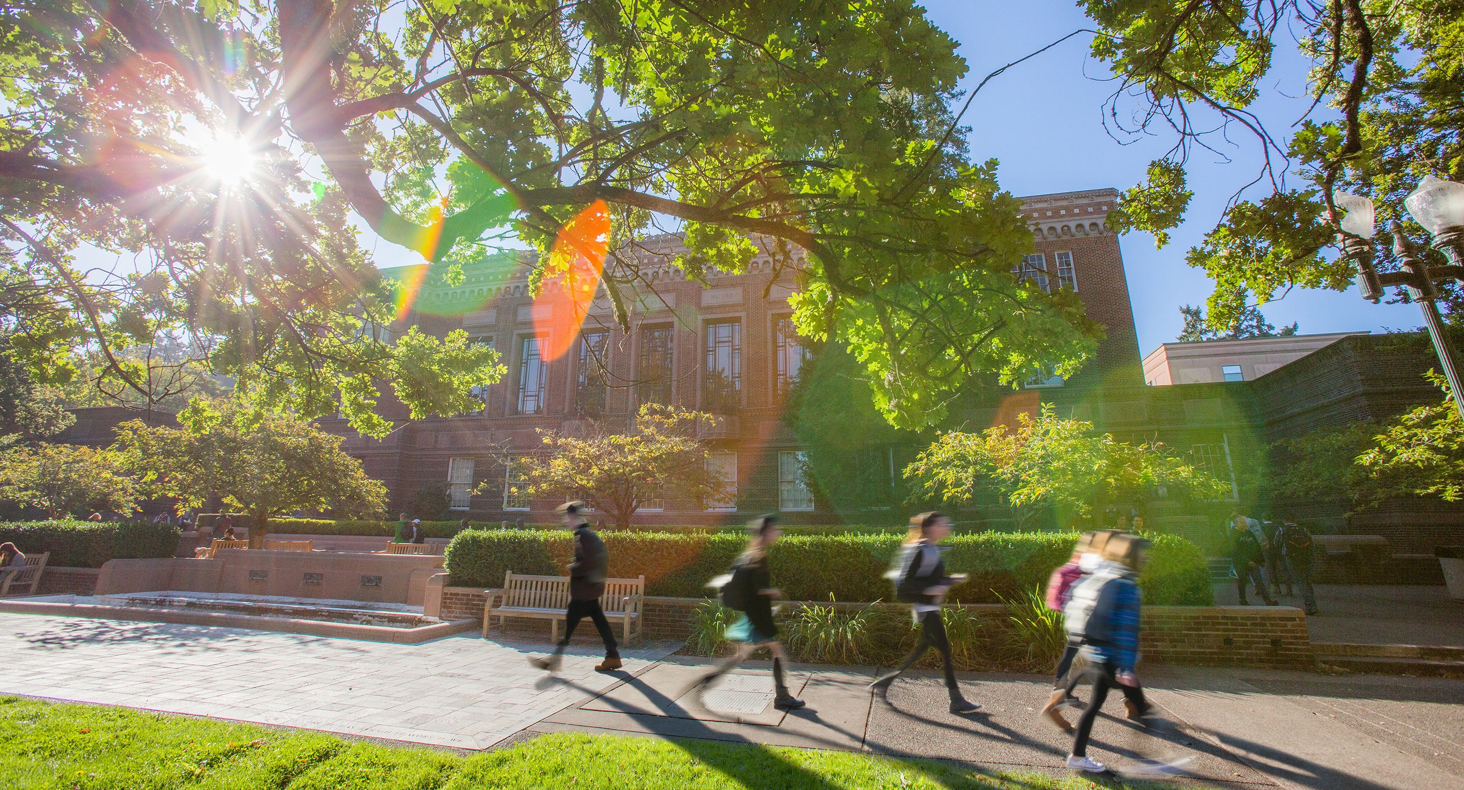 Students walking in front of the landscaped entrance for a large brick building with large ornate windows in the early morning sunlight.