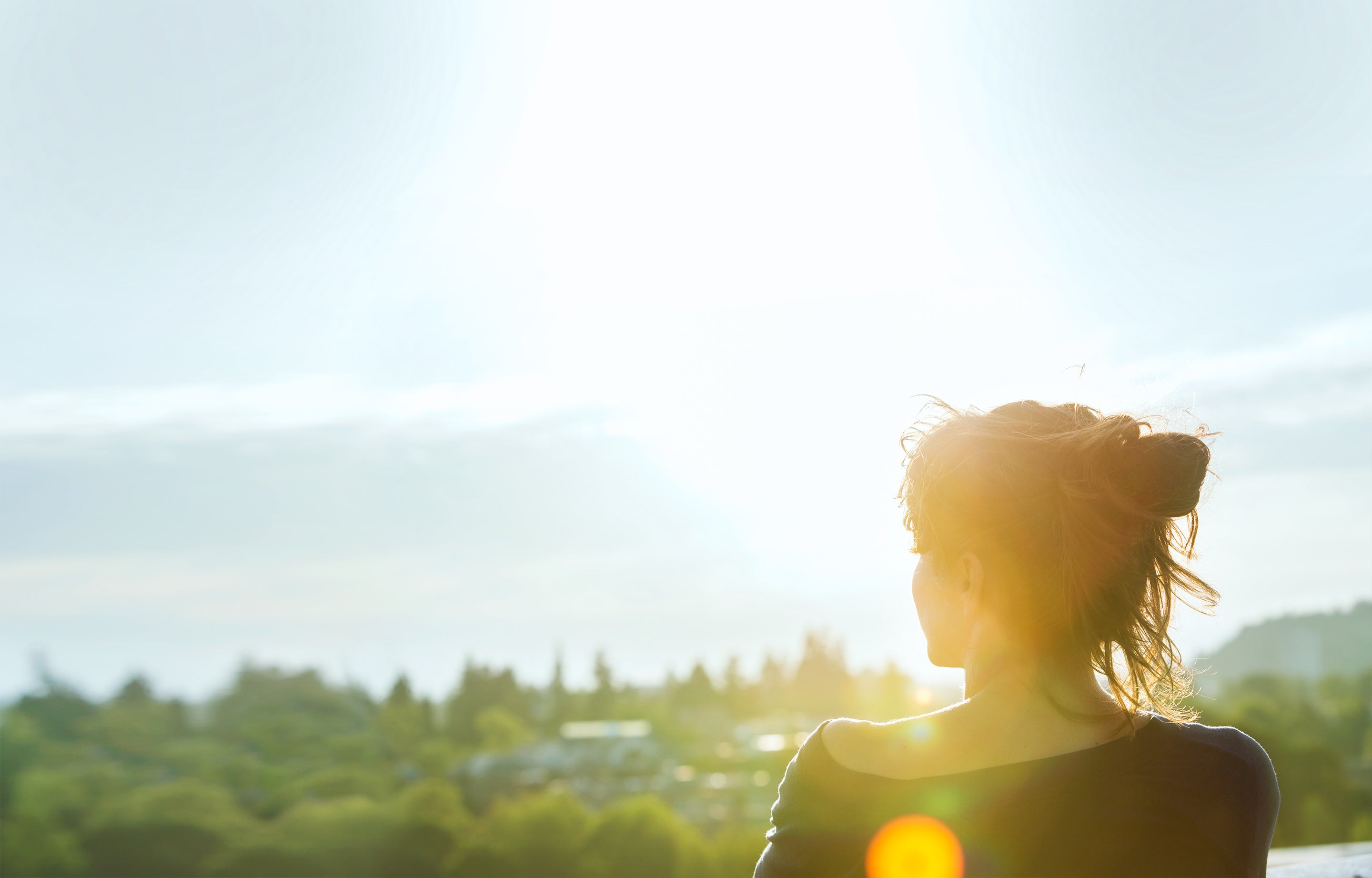 A woman on a viewpoint looking over a forested urban area in bright sunlight.