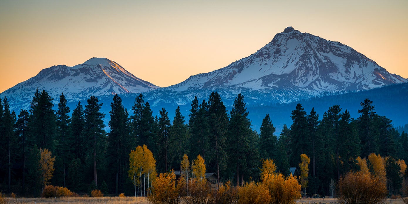 The sun sets pink and orange behind the Three Sisters Mountains, showing the snowy silhouette of the peaks rising above the fall foliage below.