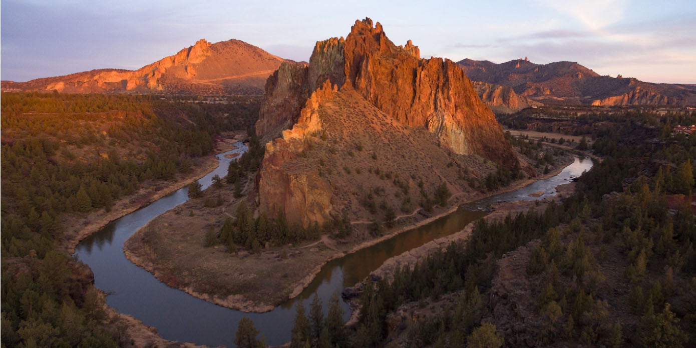 An orange sunrise illuminates the peak of Smith Rock and the surrounding mountains while the Crooked River winds below in Central Oregon's high desert.