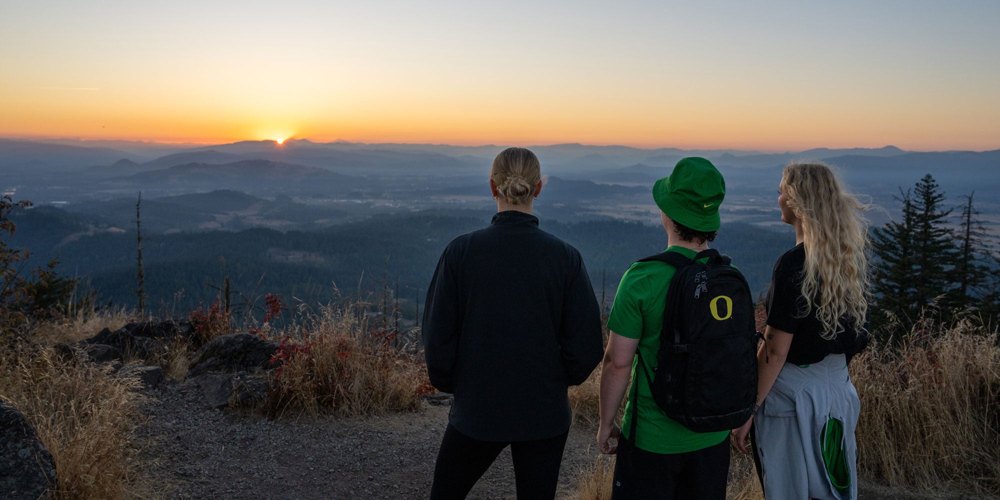 Three students looking west from a mountain top over hills and valleys towards the sunset