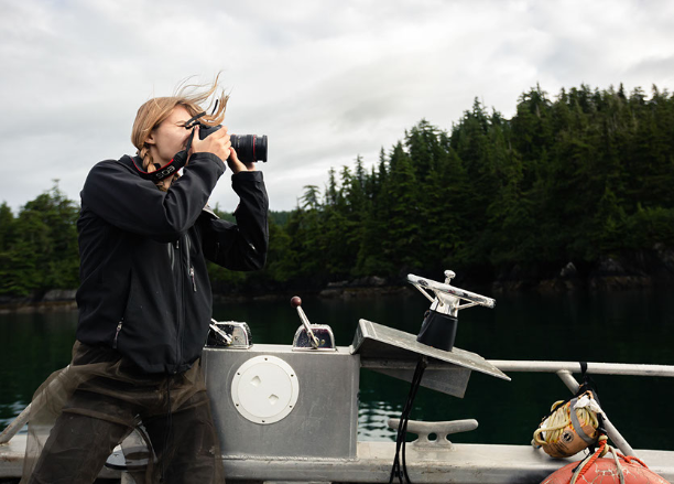 Student using a camera while standing on their boat on a lake surrounded by forest